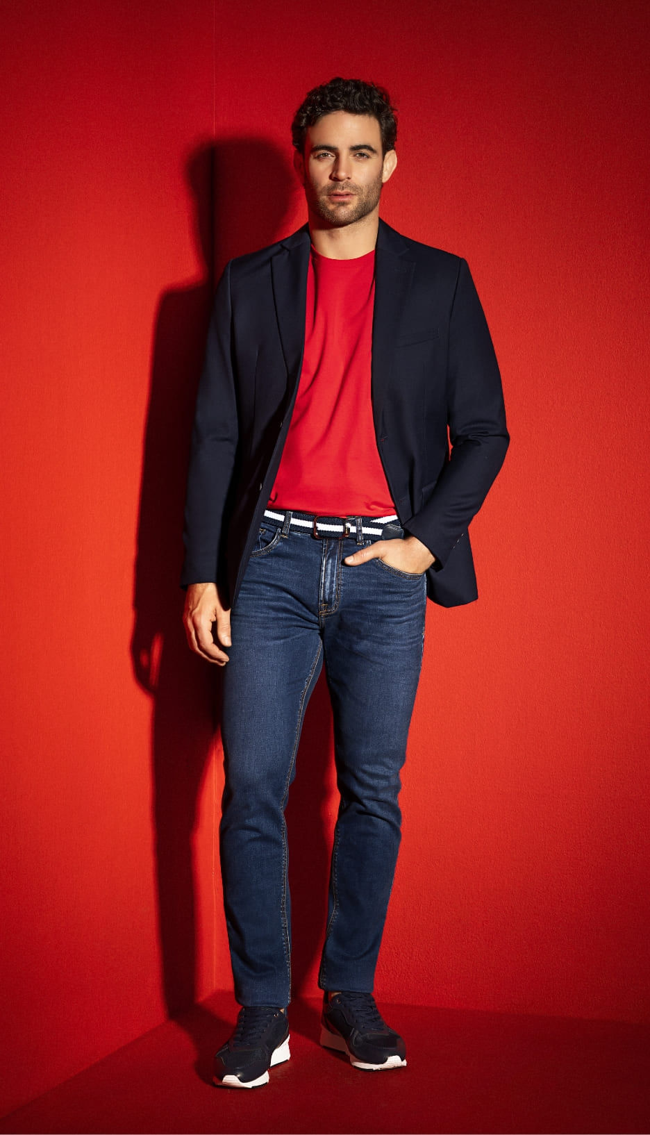Hombre con blazer azul oscuro, camiseta roja, jean skinny con cinturón azul y blanco y tenis negros de la marca Studio F MAN